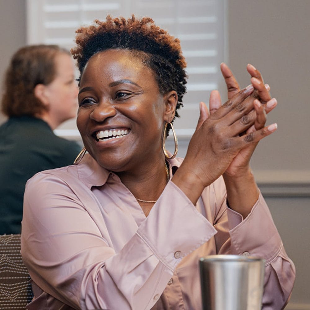A woman with short curly hair smiles and claps her hands while sitting at a table. She is wearing a light pink blouse and hoop earrings. Another person is blurred in the background.