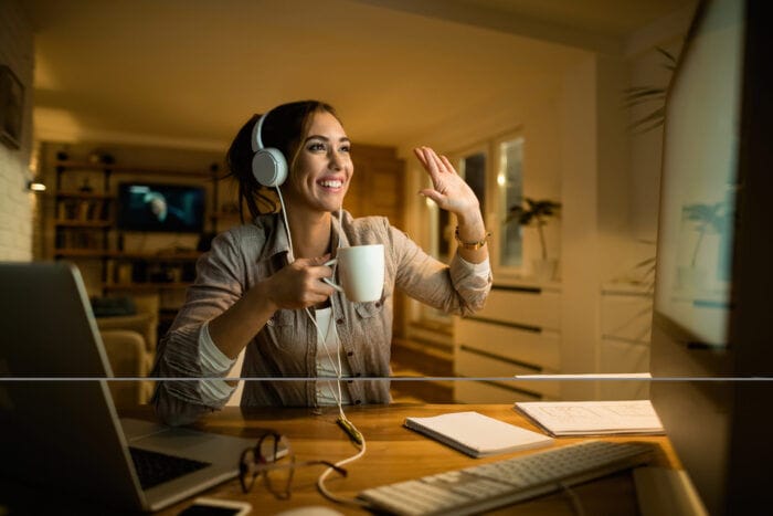 A woman with headphones on sips tea and waves hello to people in a video meeting.