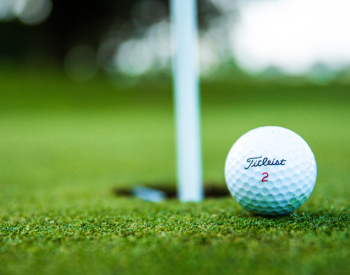 Close-up of a white golf ball labeled Titleist 2 resting on well-manicured green grass near a hole with a white flagstick. The background is softly blurred, emphasizing the ball and hole in a serene golf course setting.