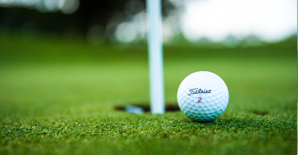 Close-up of a white golf ball labeled Titleist 2 resting on well-manicured green grass near a hole with a white flagstick. The background is softly blurred, emphasizing the ball and hole in a serene golf course setting.