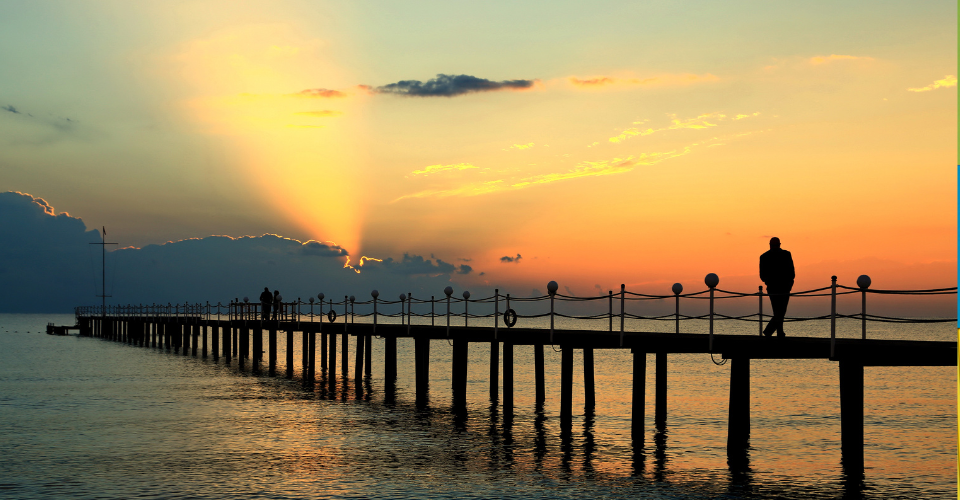 A long pier extends over calm water at sunset, with a person standing at the end. The sky is orange and yellow with scattered clouds. Sunlight beams radiate from behind the clouds, creating a dramatic effect.