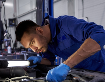 A mechanic in a blue jumpsuit and gloves is focused on working under the hood of a car. He is holding a bright inspection light to illuminate the engine area in a garage setting.