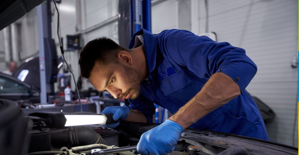 A mechanic in a blue jumpsuit and gloves is focused on working under the hood of a car. He is holding a bright inspection light to illuminate the engine area in a garage setting.