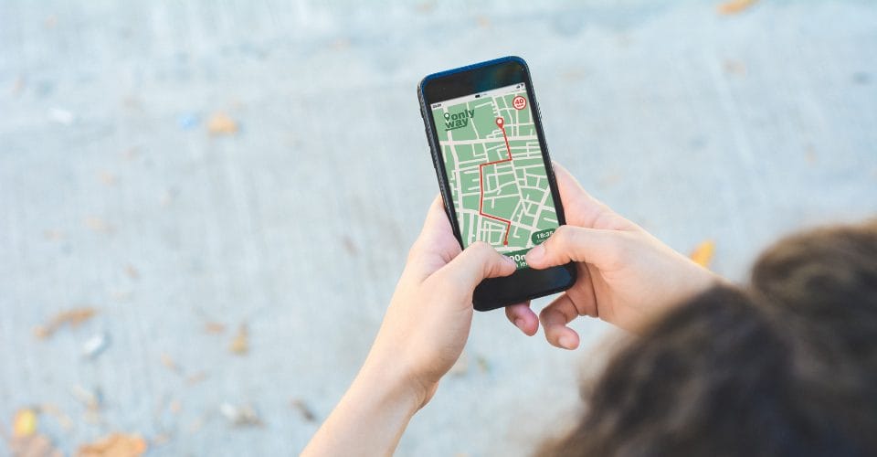 A person holds a smartphone displaying a digital map with a marked route. The screen shows detailed streets on a green background. The phone is held with both hands against a gray pavement with scattered leaves in the background.