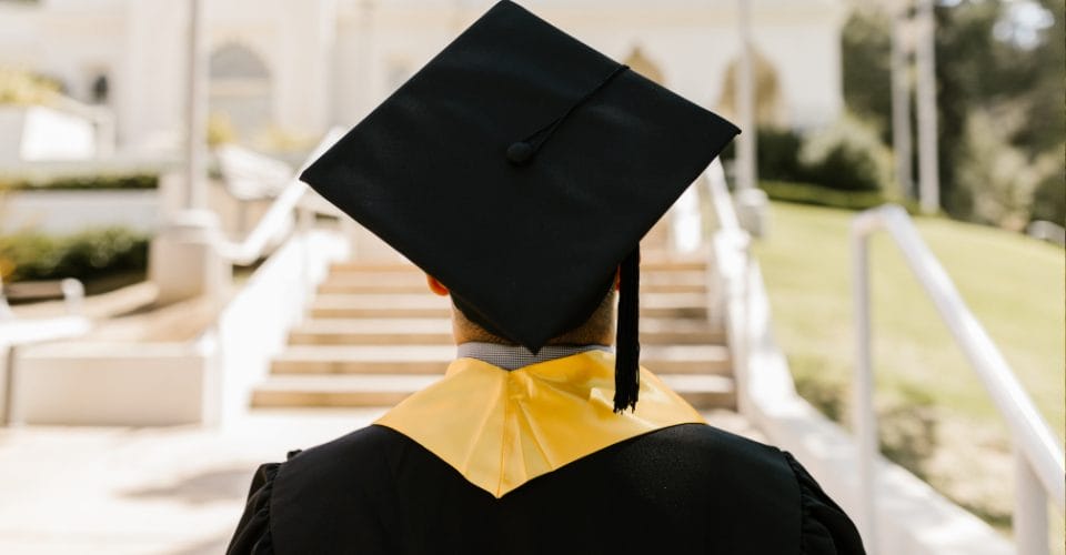 A graduate in a black cap and gown with a yellow stole stands facing a set of stairs outdoors. The background is blurred, showing greenery and part of a building. The focus is on the back of the graduate.