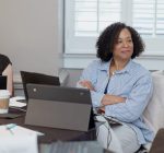 Two women sit at a table with laptops, notebooks, and coffee, attentively listening during a nonprofit or higher ed technology consulting meeting in a bright room with a window and shutters in the background.