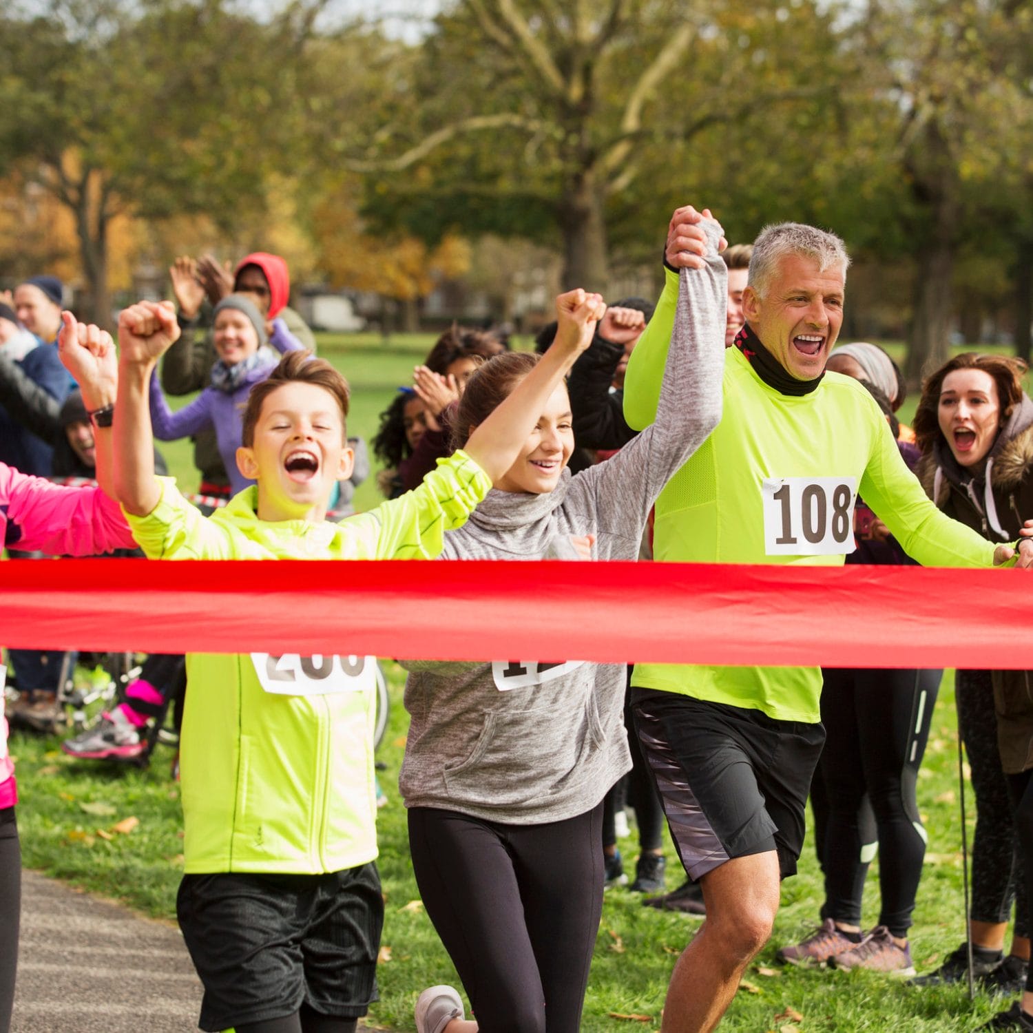 A group of runners cheer as they cross a red finish line in a park, holding hands in victory, while a crowd of spectators celebrates in the background.