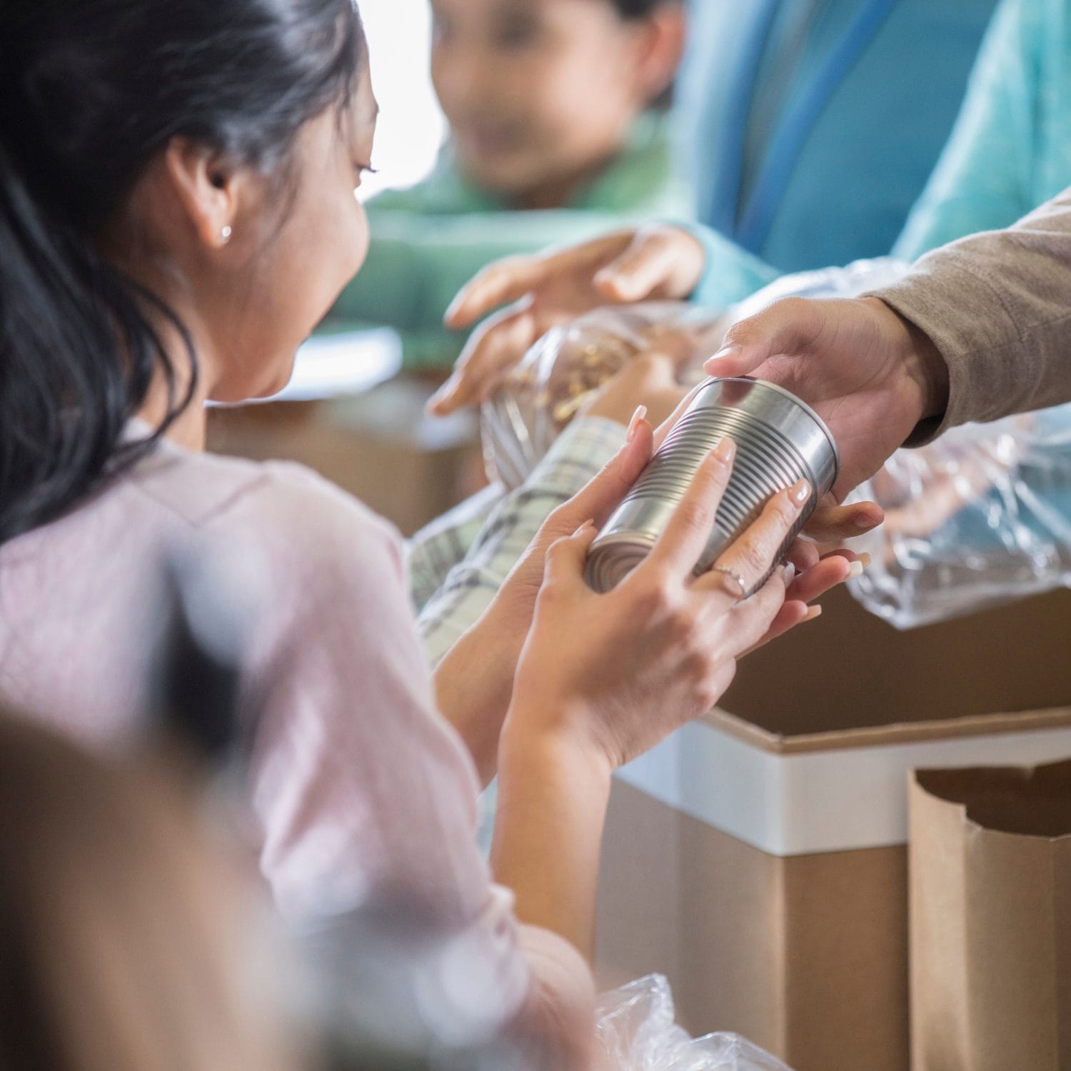 A woman receives a canned food item from another person at a food donation or distribution event. Cardboard boxes and other people are visible in the background.