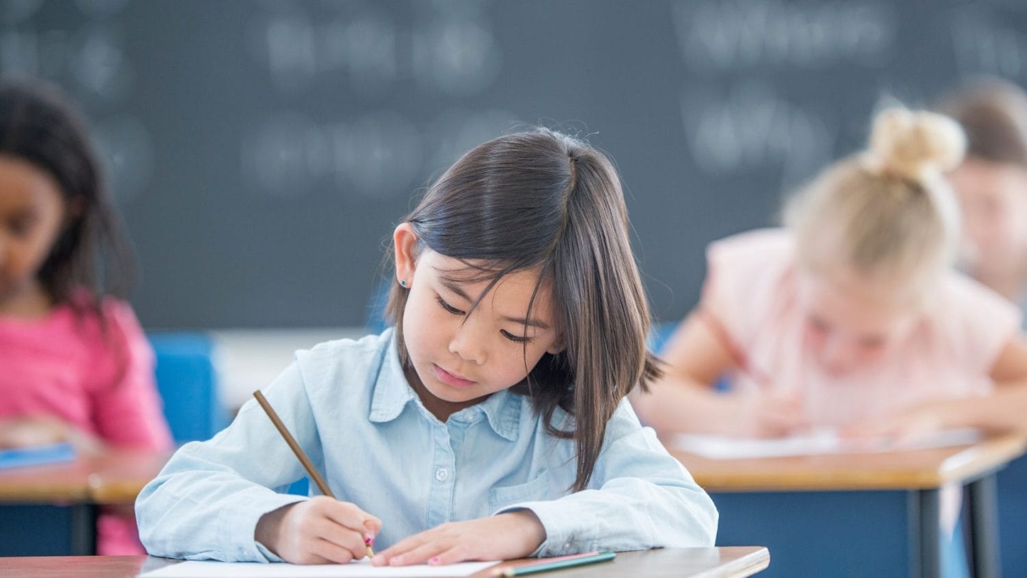 A young girl sits at a classroom desk, focused on writing in her notebook. Other children are also working in the background, and a chalkboard is visible behind them.