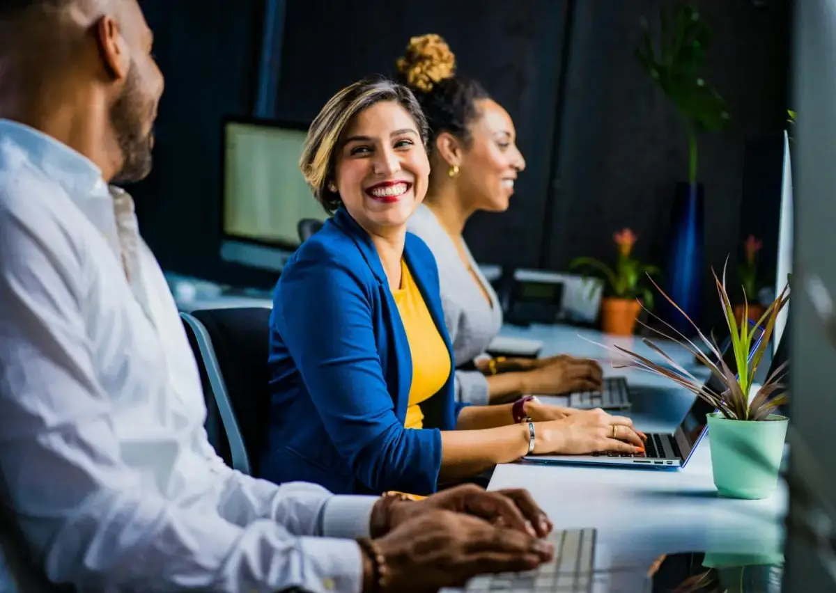 Three people sit at a desk working on laptops in an office. The woman in the center, wearing a blue blazer and yellow top, smiles brightly at the camera. The other two people are focused on their work.