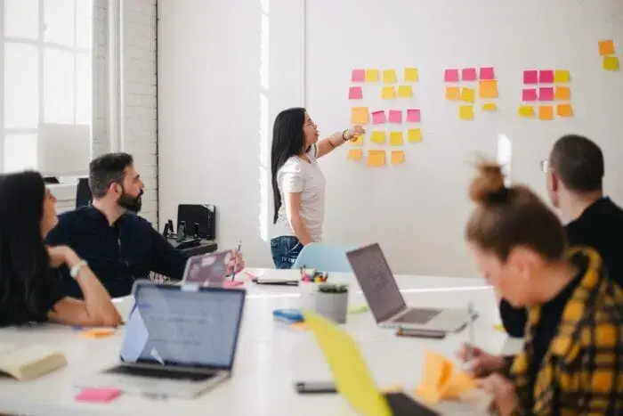 A woman stands by a wall covered with colorful sticky notes, pointing at one while four colleagues sit at a table with laptops, listening and taking notes in a bright, modern office.