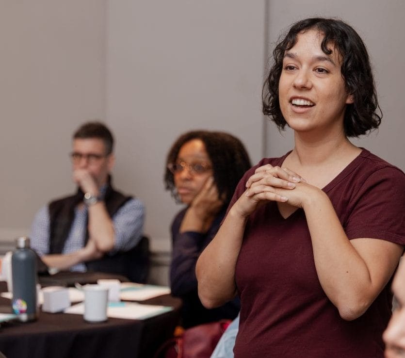 A woman stands and speaks with her hands clasped, smiling, in a room where others listen attentively. Tables with papers, cups, and water bottles suggest a workshop on Kindsight Fundraising Platform ascend implementation and optimization services.