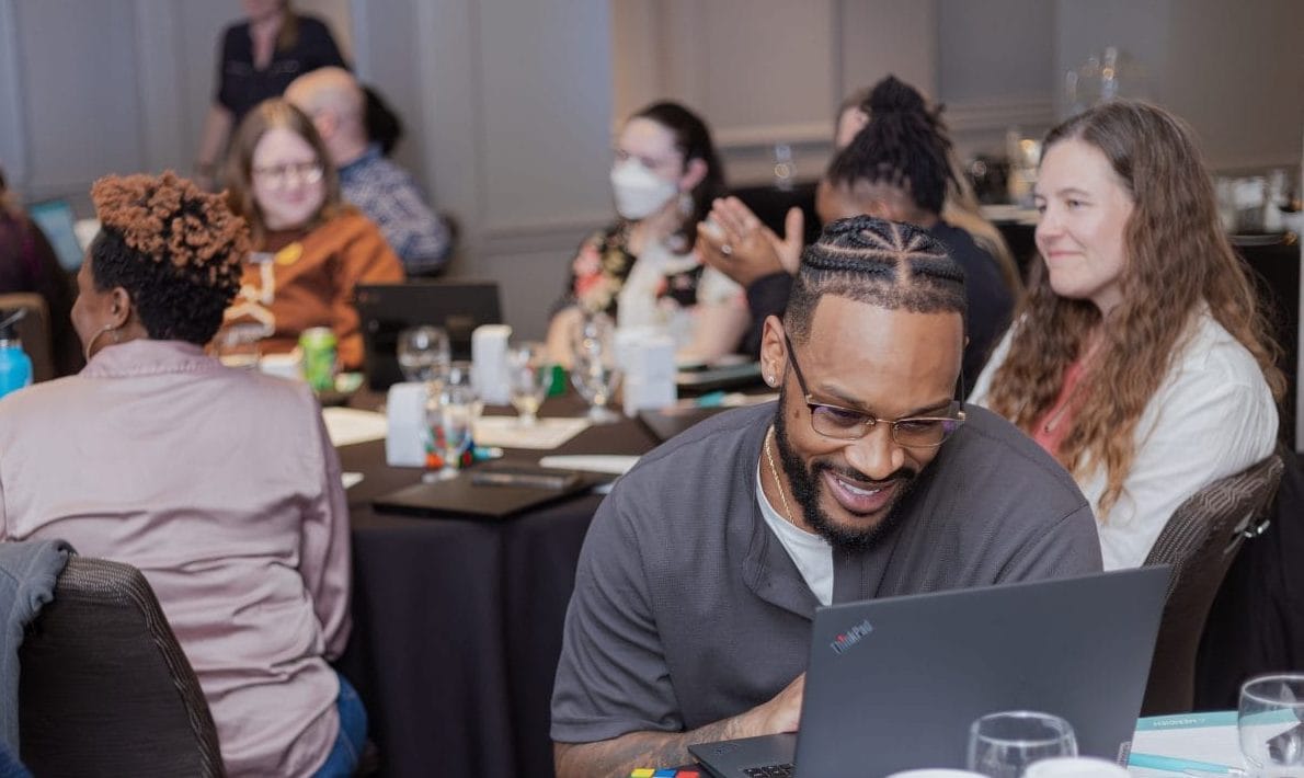 A group of people sit at round tables during a meeting or workshop. One man in the foreground smiles while working on a laptop. Others are talking, laughing, or listening, creating a friendly, engaged atmosphere.