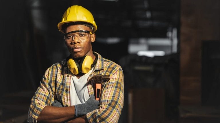 A construction worker wearing a yellow hard hat, safety glasses, ear protection, and gloves stands with arms crossed, holding a hammer, in a dimly lit industrial setting.