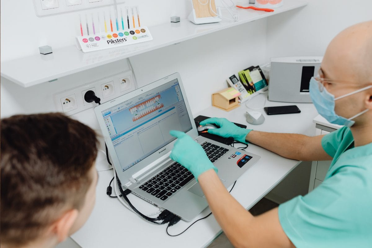 A dentist wearing gloves and a face mask explains dental data on a laptop to a patient in a modern dental clinic. Dental models and tools are visible on the desk and shelves.