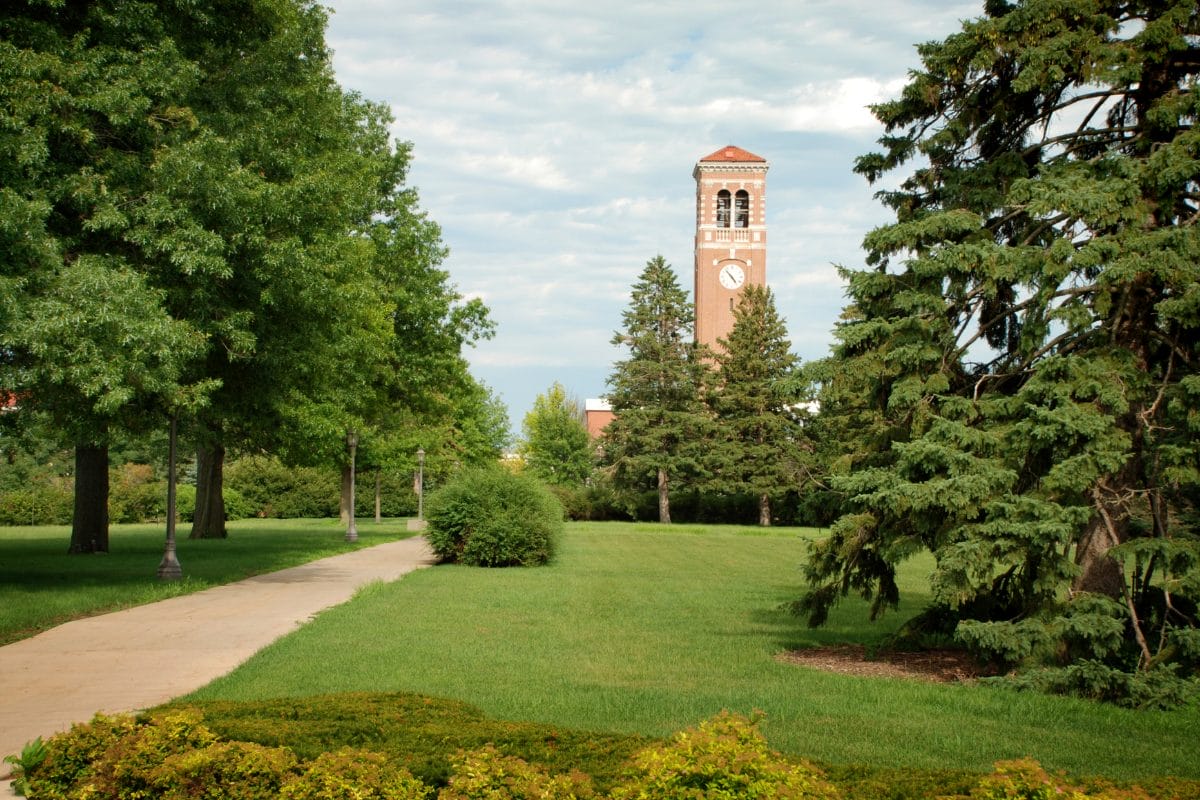 A paved path curves through a green, grassy campus with tall trees. In the background, a brick clock tower rises above the trees under a partly cloudy sky.