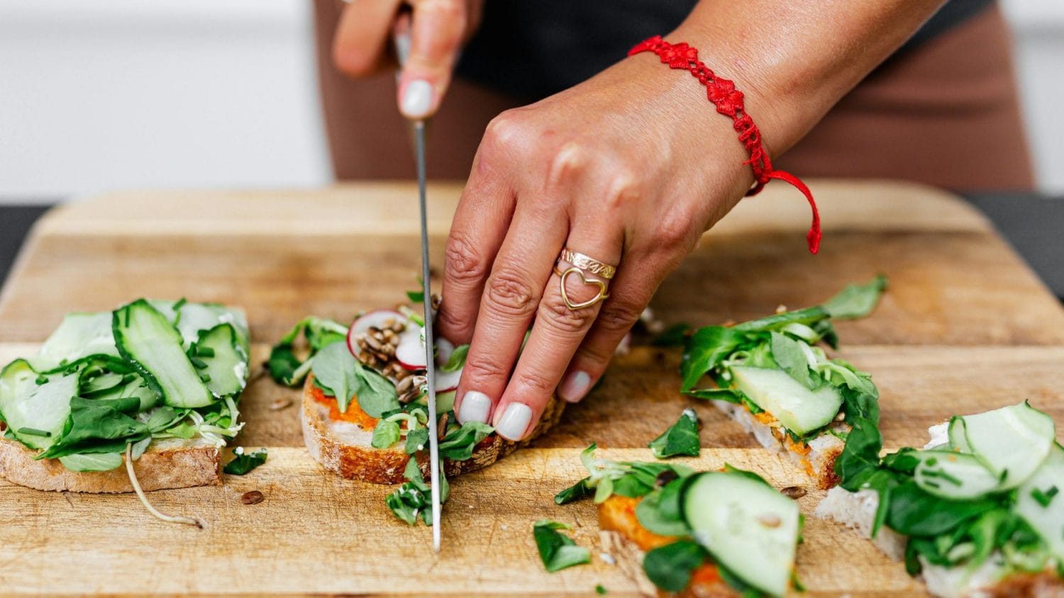 A person cuts slices of bread topped with greens, cucumber, and other vegetables on a wooden cutting board. The person wears a red bracelet and rings.