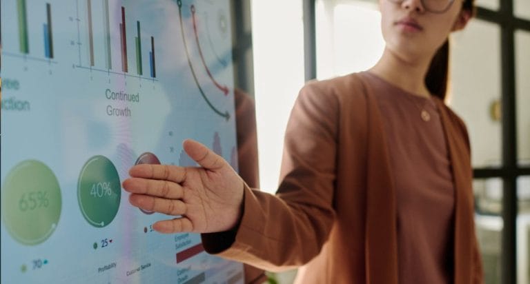 A person in business attire points at a large digital screen displaying colorful nonprofit data management graphs, charts, and analytics data, including pie charts with percentages and bar graphs, in a modern office setting.