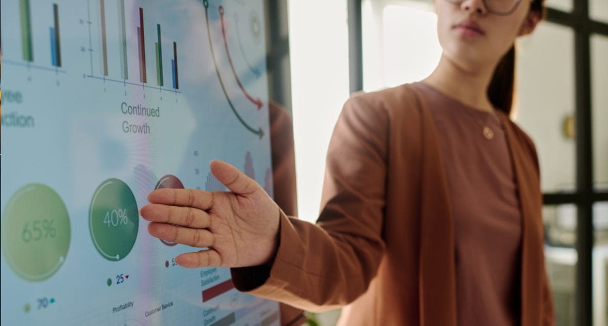 A person in business attire points at a large digital screen displaying colorful nonprofit data management graphs, charts, and analytics data, including pie charts with percentages and bar graphs, in a modern office setting.