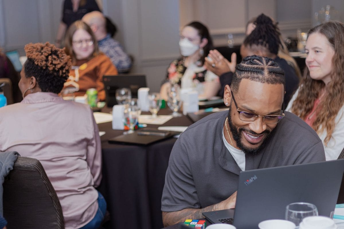 A group of people sit at tables with laptops during a meeting or workshop. One man in the foreground smiles at his laptop, while others in the background are talking or listening. Some people are wearing masks.