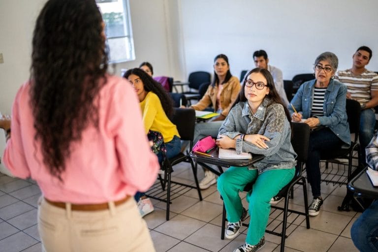 A group of students sitting at desks listen attentively to a teacher standing at the front of a classroom. One student in green pants sits facing forward, taking notes. The room is bright with natural light.
