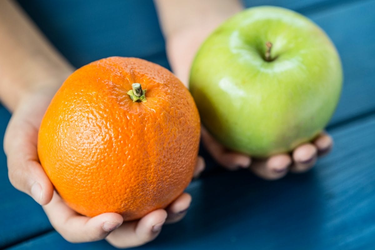 A person is holding an orange in one hand and a green apple in the other, against a blue wooden background.