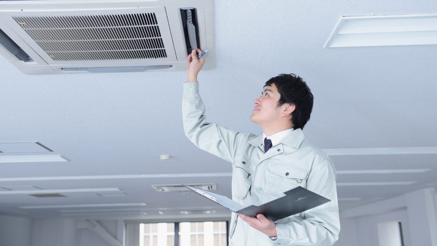 A technician in a light-colored uniform inspects and writes on a ceiling-mounted air conditioning unit with a clipboard, much like outlining an AI roadmap for nonprofits case study, inside a modern office space.