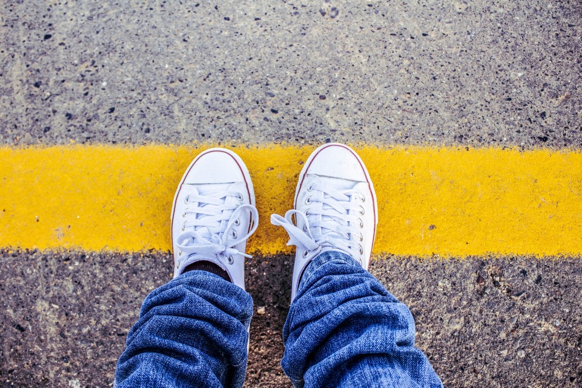 A person wearing white sneakers and blue jeans stands at a yellow line painted on a rough concrete surface, viewed from above.