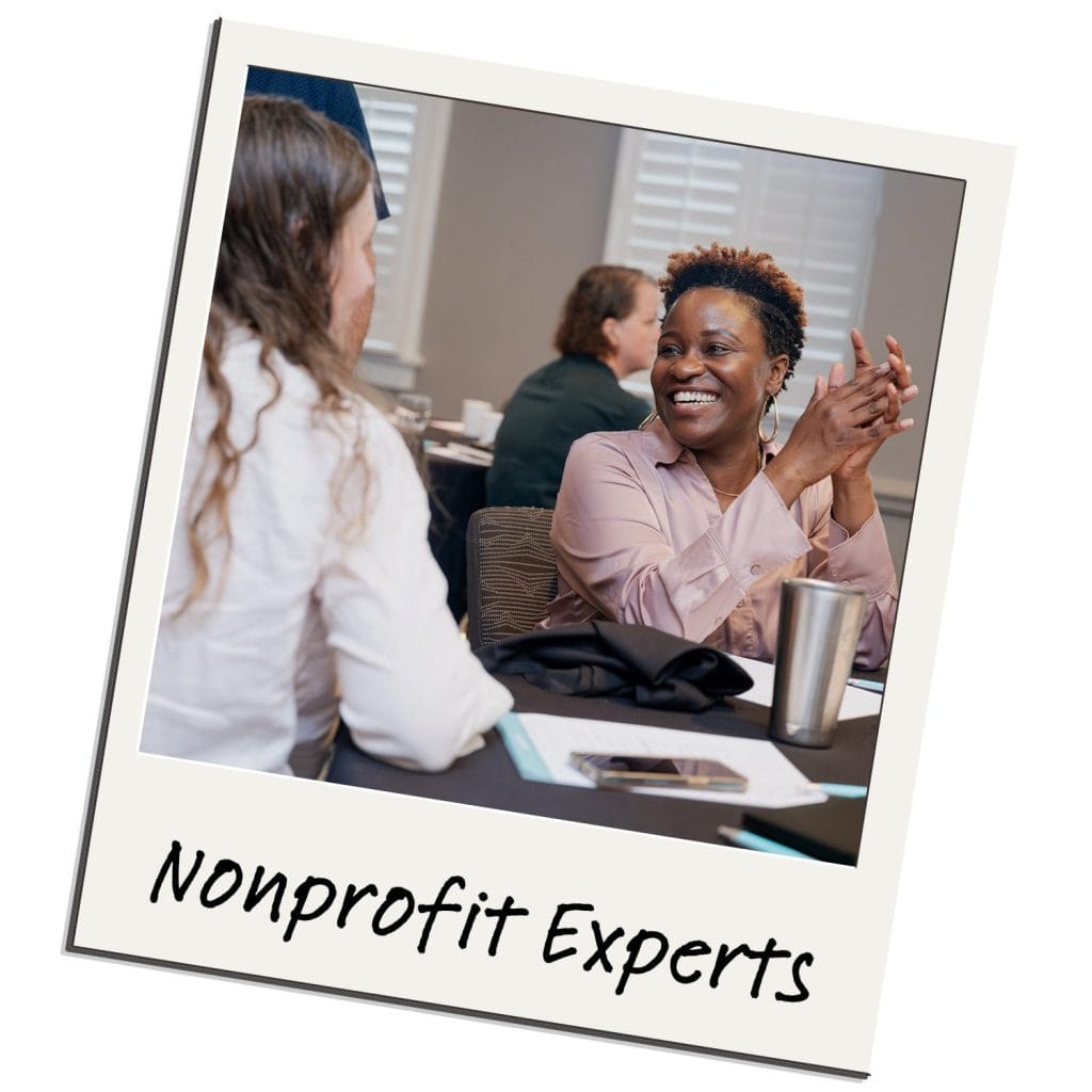 Two women sit at a table in conversation during a meeting or workshop. One woman is smiling and clapping. Notebooks, paper, and a tumbler are on the table. The text at the bottom reads “Nonprofit Experts—powered by Heller Consulting Donor Data Hub.”.