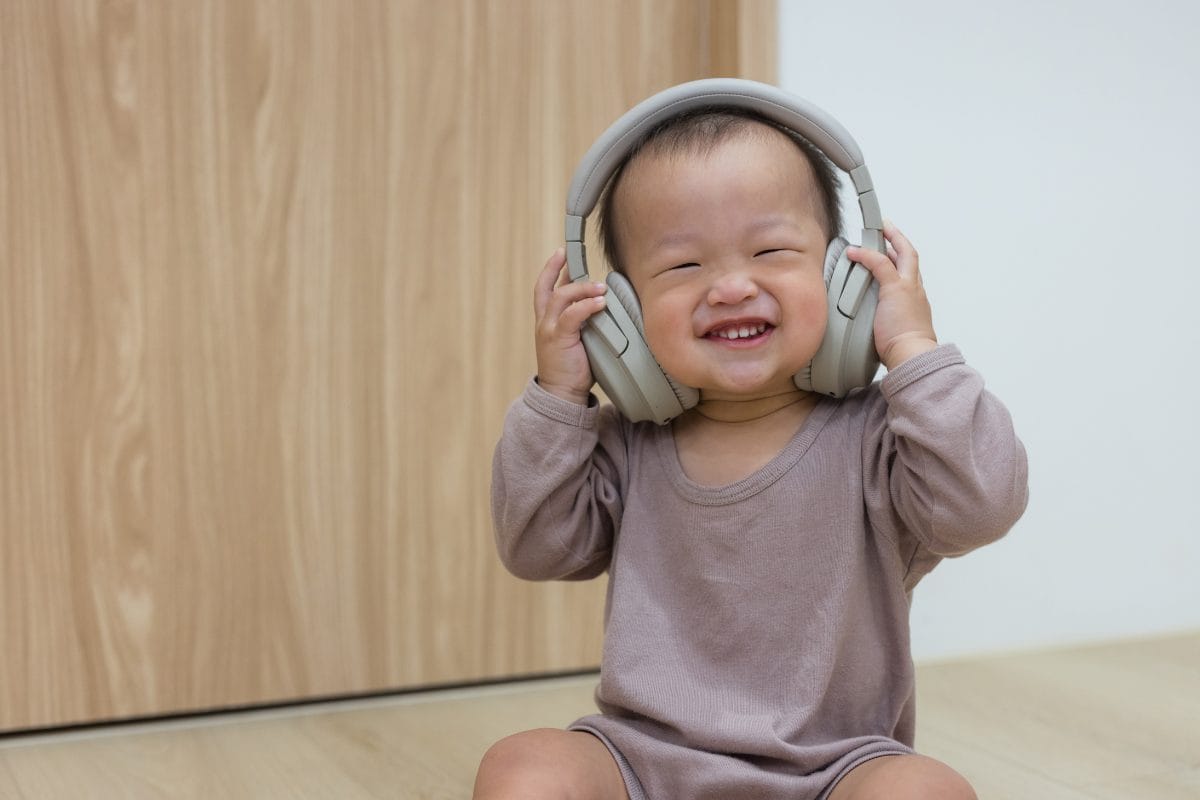 A smiling baby wearing a long-sleeved, light brown outfit sits on a wooden floor, holding large over-ear headphones to their head, looking happy and playful.