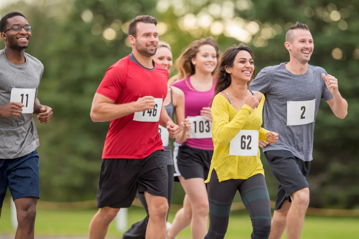 A group of smiling runners wearing race numbers jog together outdoors on a path, surrounded by greenery. The atmosphere appears cheerful and energetic.