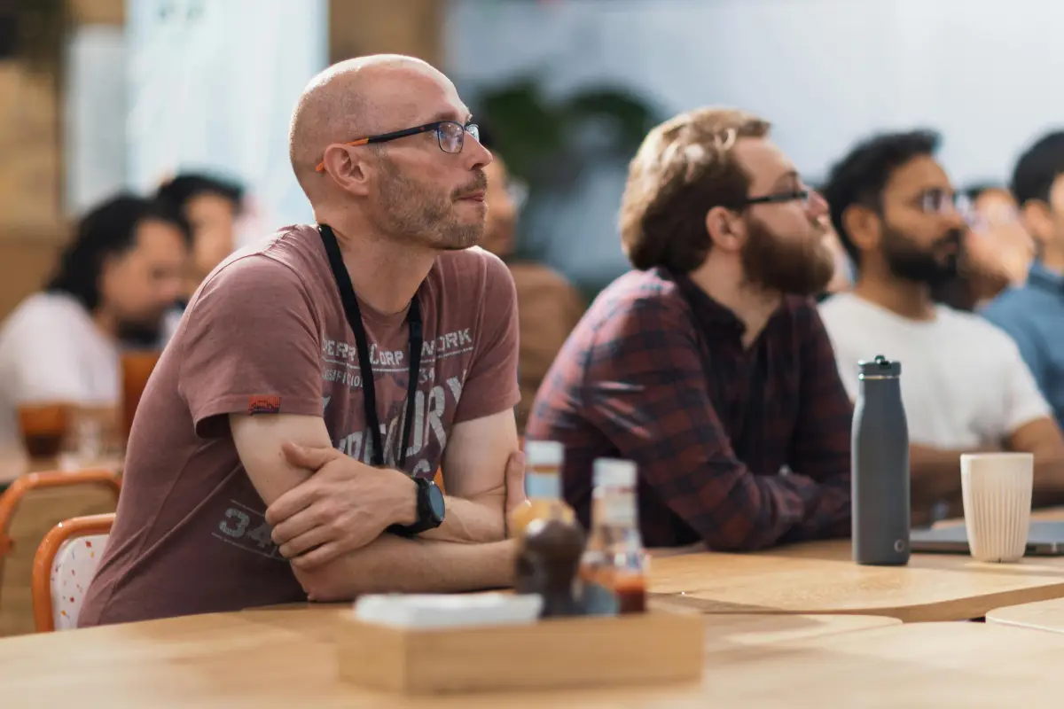 A group of people sit at a table, attentively listening to something off-camera. The focus is on a bald man with glasses and a maroon t-shirt, with others beside him. Bottles and a cup are on the table.