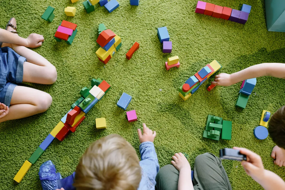 Three children sit on a green carpet, playing with colorful wooden building blocks scattered around them. Their faces are not visible, and they are focused on constructing various shapes and structures.