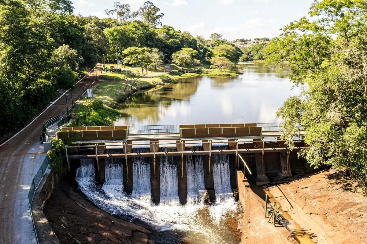 A small dam with water flowing over its gates sits on a river surrounded by lush green trees and a dirt road running parallel to the riverbank under a bright, sunny sky.