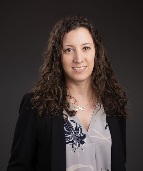 A woman with long curly brown hair, wearing a dark blazer over a floral blouse, stands against a plain dark background and smiles at the camera.