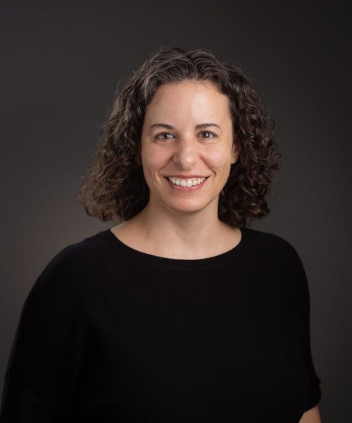 A woman with curly brown hair wearing a black top smiles at the camera against a plain dark background. Her left hand is in her pocket.