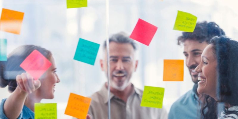 A group of four people smiling and discussing around a glass wall covered with colorful sticky notes, reflecting their collaborative spirit in nonprofit work. The notes contain various handwritten words, showcasing innovative ideas in higher ed technology consulting. The atmosphere is vibrant and positive.