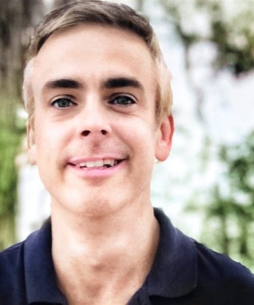 A young man with short blonde hair, wearing a dark polo shirt, smiles at the camera while standing outdoors with trees and greenery in the background, exuding the approachable confidence of a skilled nonprofit consultant.