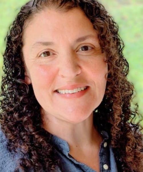 A woman with curly brown hair smiles at the camera. She is wearing a blue textured shirt and stands in front of a blurred green outdoor background, embodying the confidence of a Kevin Hartland nonprofit data expert.