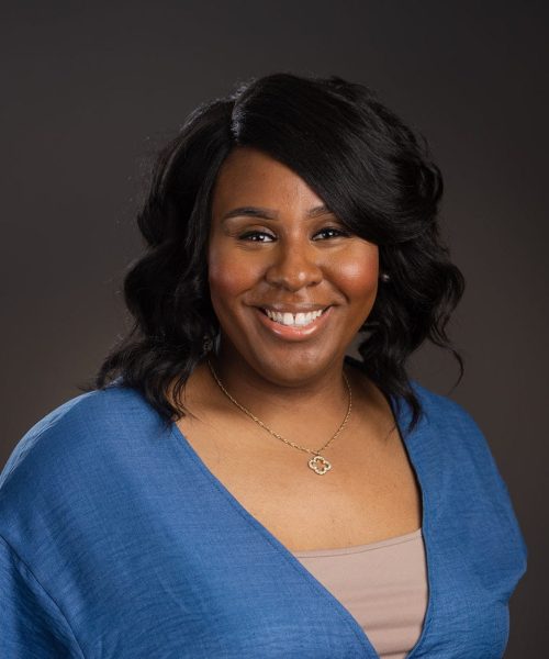 A woman with wavy black hair smiles at the camera. She is wearing a blue wrap top over a beige shirt and a flower-shaped necklace, standing in front of a plain dark background.