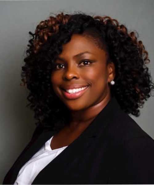 A woman with curly black and brown hair smiles at the camera. She is wearing a black blazer over a white shirt, with pearl earrings, and is posed in front of a plain gray background.