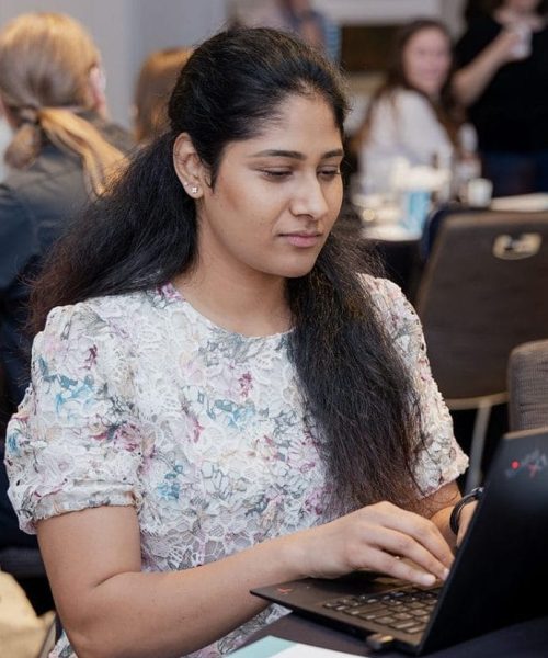 Heller Consulting developer Manasa Kamepally, a woman with long dark hair in a floral blouse, works on her laptop at a meeting table. Other people are talking in the background, with notebooks and coffee cups scattered on the table.