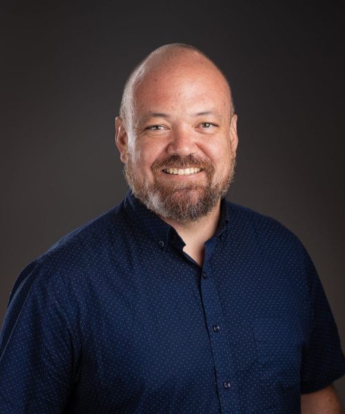 A smiling man with a bald head and beard, wearing a short-sleeved navy blue button-up shirt with small dots, stands in front of a dark, plain background.