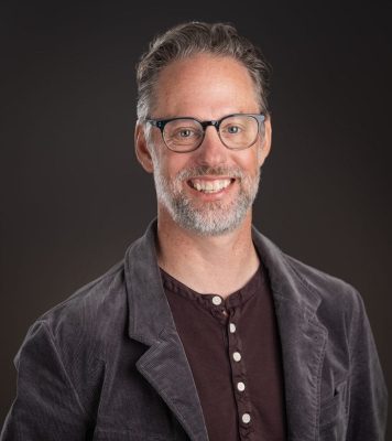 A smiling man with short, gray hair and a beard, dressed as a nonprofit consultant in glasses, a dark buttoned shirt, and a gray corduroy jacket, stands against a plain dark background.