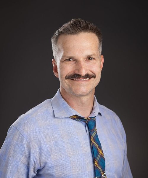 A man with short brown hair and a mustache, wearing a light blue checkered shirt and a plaid tie, smiles at the camera against a dark background.