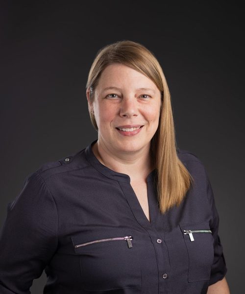A woman with straight, light brown hair, wearing a dark long-sleeve shirt with zippered pockets, stands smiling with her hands in her pockets against a dark, plain background.