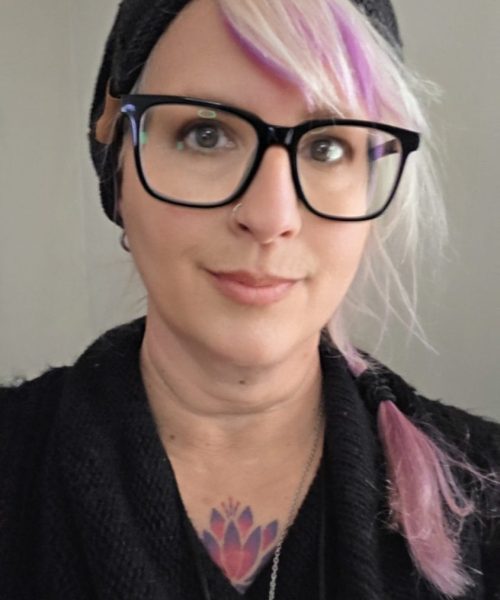 A nonprofit consultant with light skin, wearing black glasses and a black beanie, has light pink and purple hair styled in a side braid. They are dressed in a black sweater and have a colorful lotus tattoo on their chest.