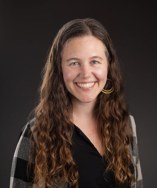 A woman with long, wavy brown hair smiles at the camera. She is wearing a black top, a plaid cardigan, and large hoop earrings. The background is plain and dark.