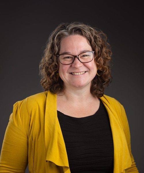 A woman with curly brown hair and glasses smiles at the camera. She is wearing a mustard yellow jacket over a black top, standing in front of a dark gray background.