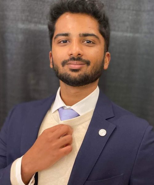 A man with short dark hair and a beard, dressed as a nonprofit consultant in a navy suit, cream sweater, white shirt, and light blue tie, stands before a dark background adjusting his tie and looking confidently at the camera.